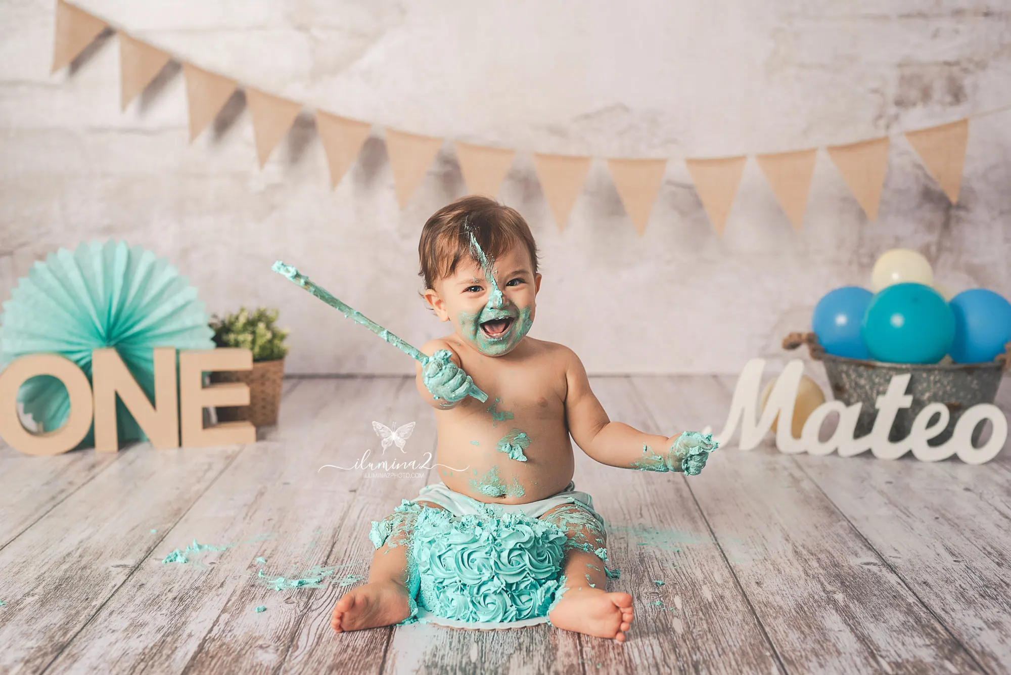 Bebé riendo con una cuchara mientras juega con su tarta de color menta en una sesión Smash Cake en Barcelona