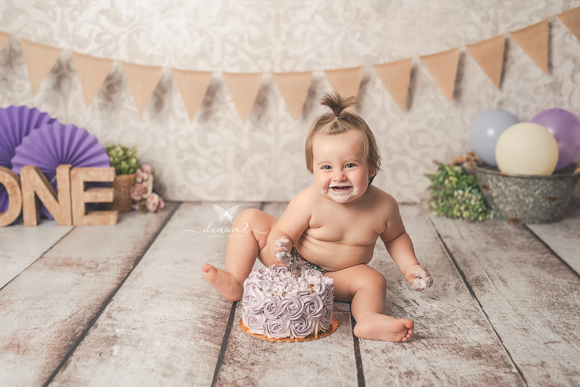 Bebé riendo junto a su pastel de color lila durante una sesión Smash Cake en estudio